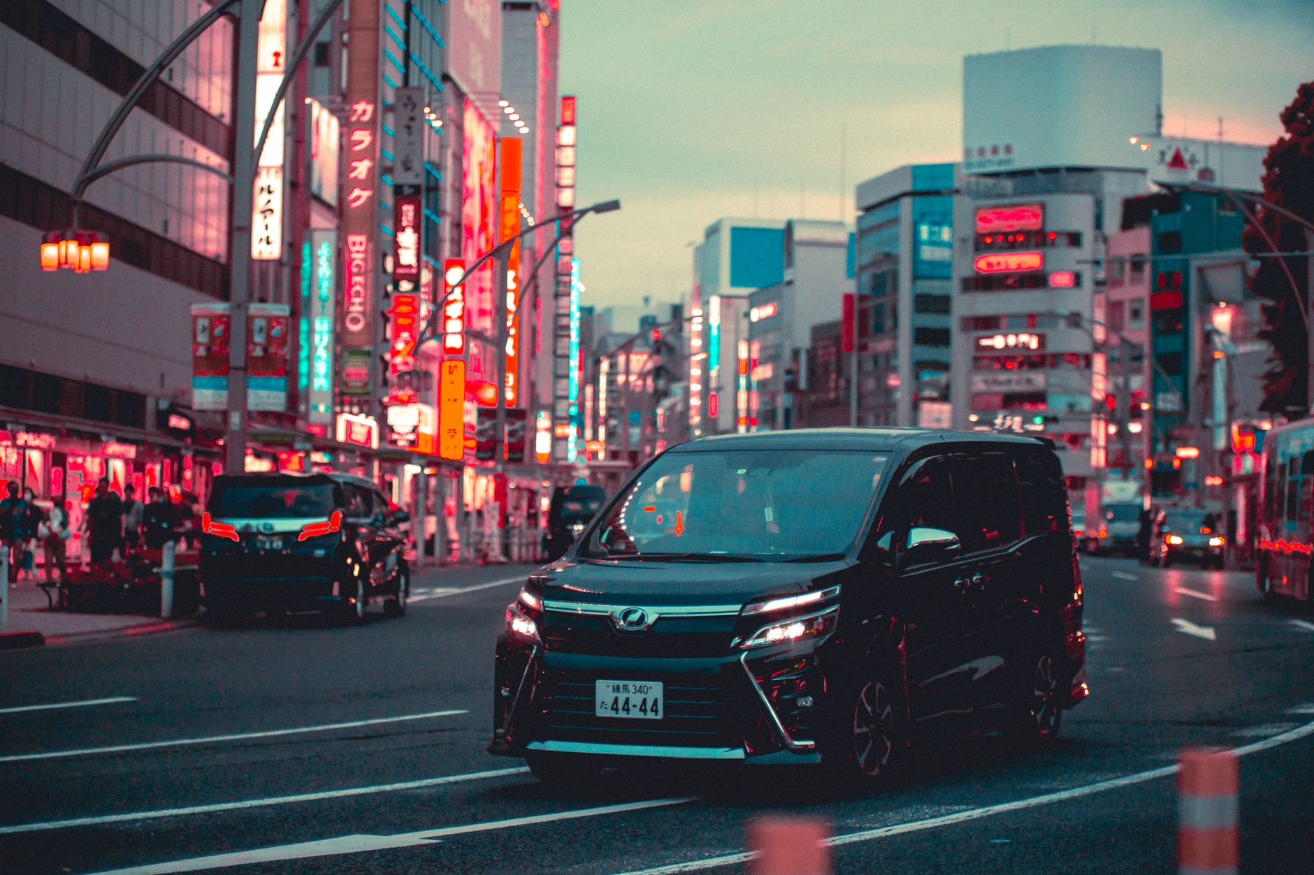 Black Japanese minivan driving on a neon-lit Tokyo street with glowing signs and other cars nearby