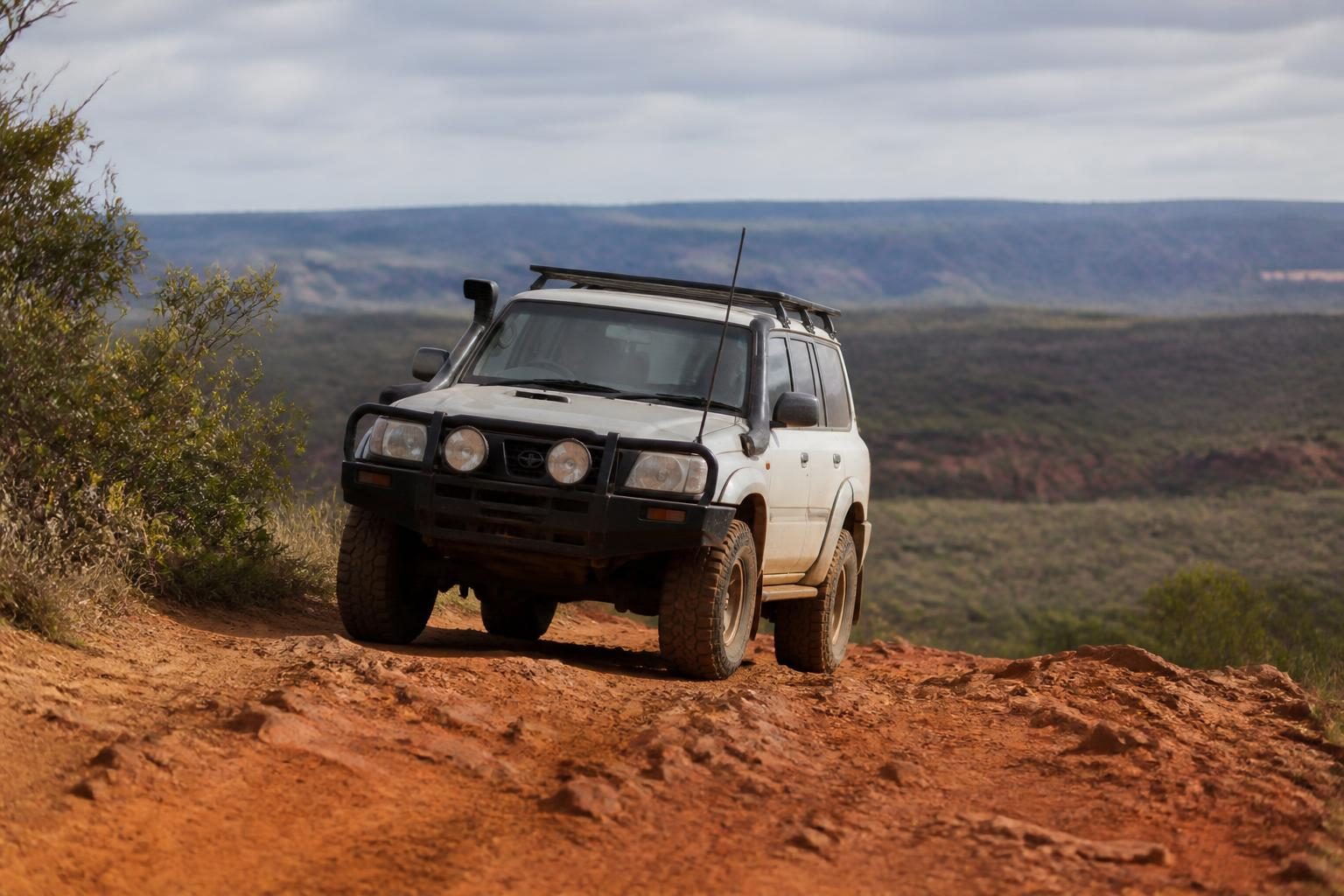 White 4WD SUV on a rocky red dirt track with expansive bushland and cloudy sky behind