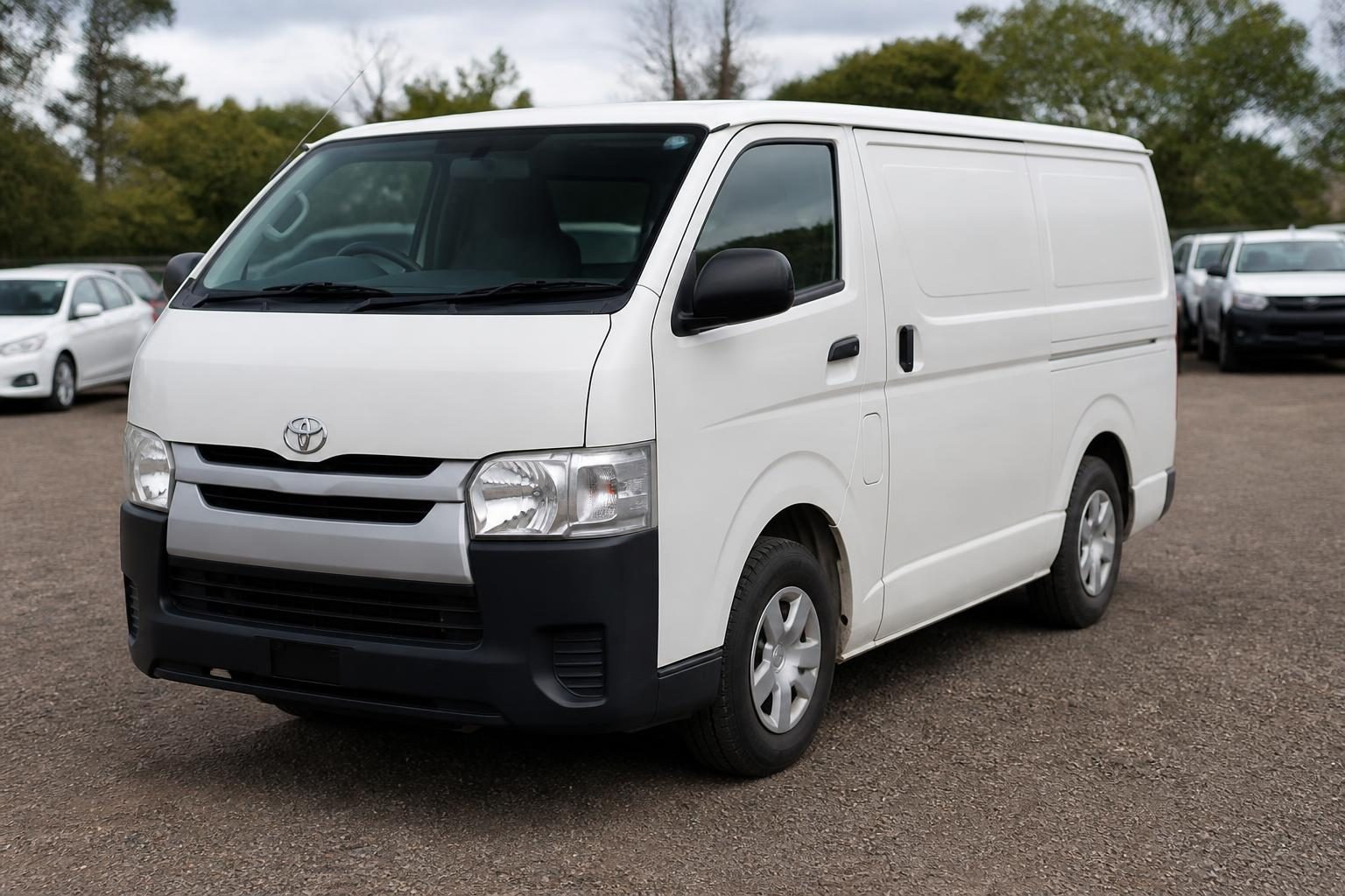 White Toyota HiAce cargo van parked on a gravel lot, shown from the front-left angle