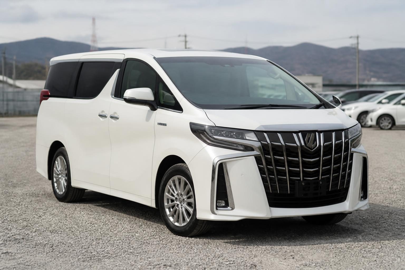 White Toyota Alphard hybrid minivan parked on a gravel lot with mountains behind