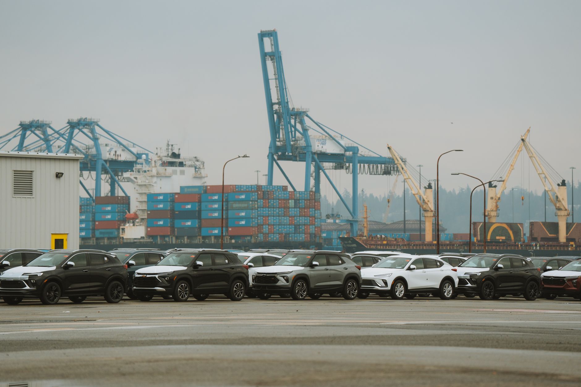 Rows of new SUVs parked at a container terminal with cranes and stacked shipping containers.