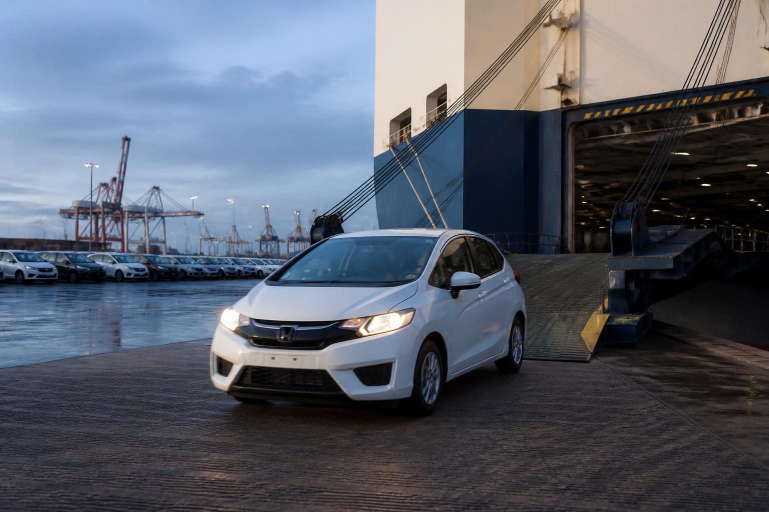 White hatchback exits a cargo ship ramp beside parked cars and container cranes at dawn.