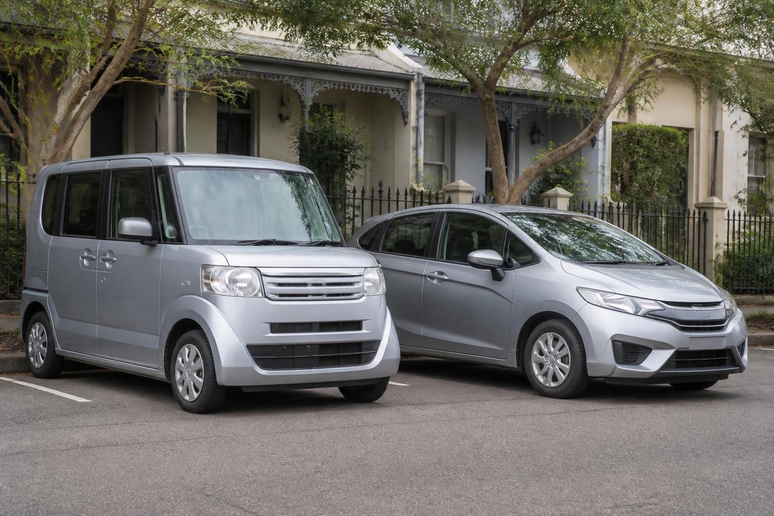 Silver boxy Honda N-Box parked beside a larger silver hatchback in front of terrace houses.