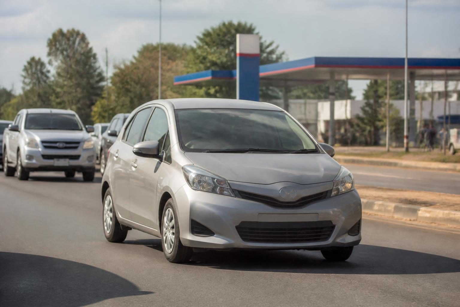 Silver compact hatchback driving on a sunlit urban road with traffic and a fuel station behind.
