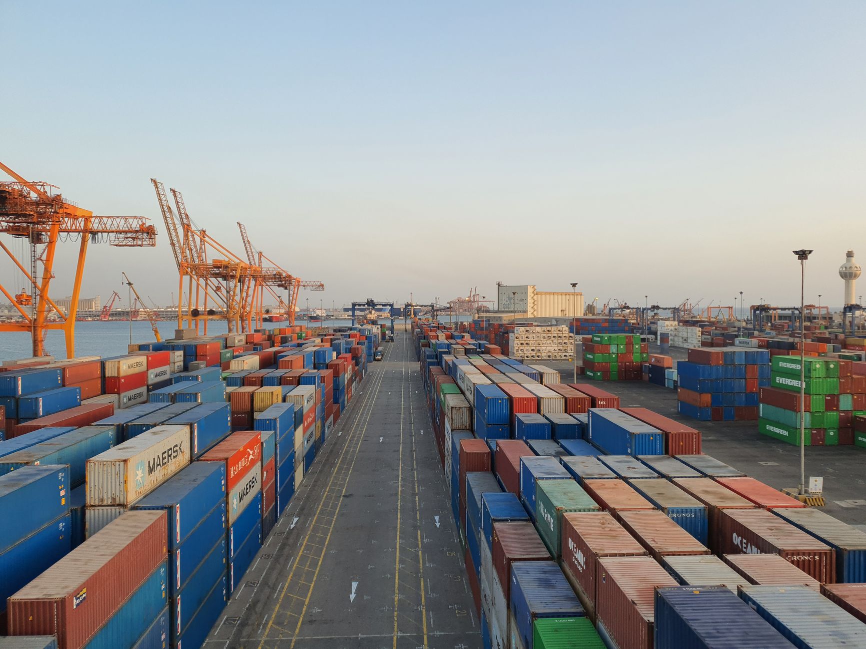 Stacks of multicolored shipping containers at a cargo port with cranes beside the water