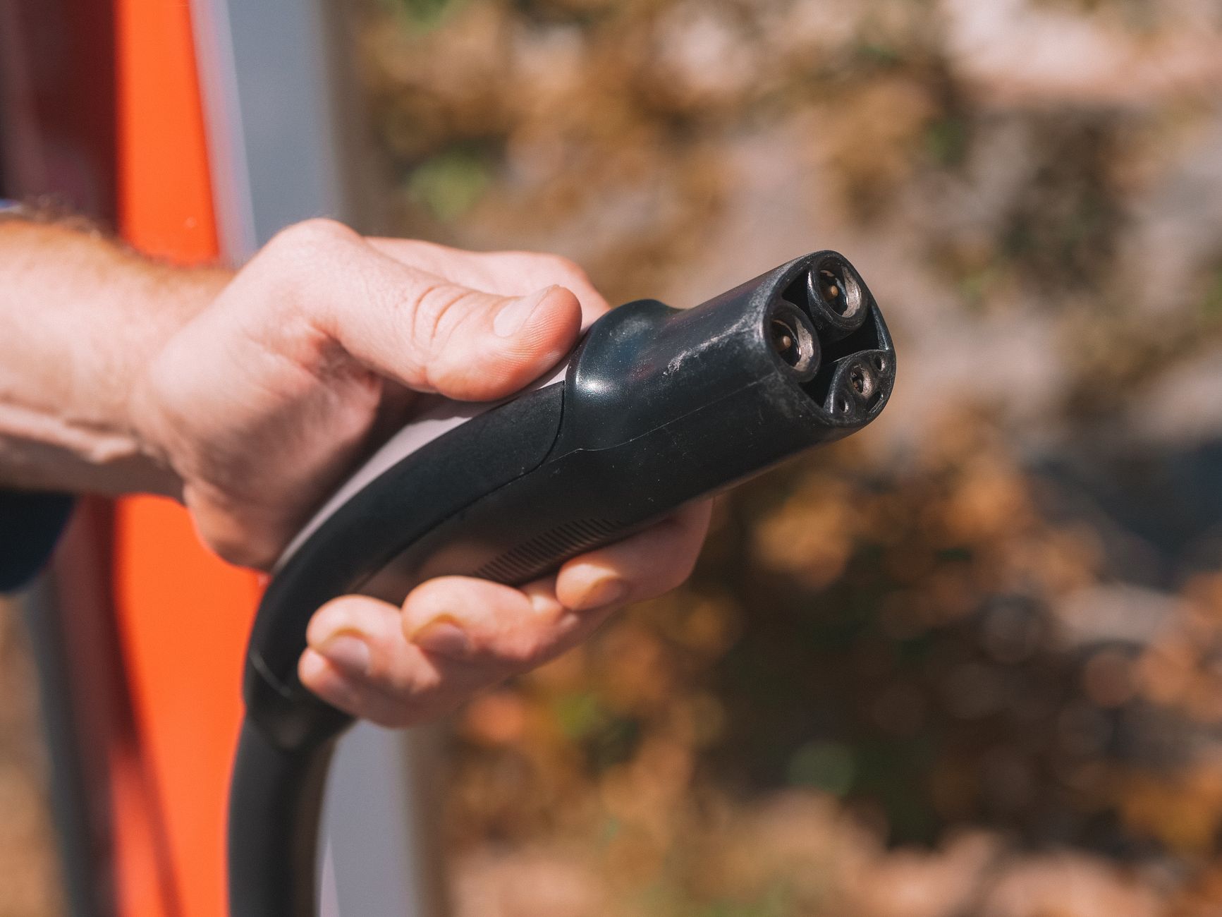 Hand holding a black electric vehicle charging connector beside a red charging station