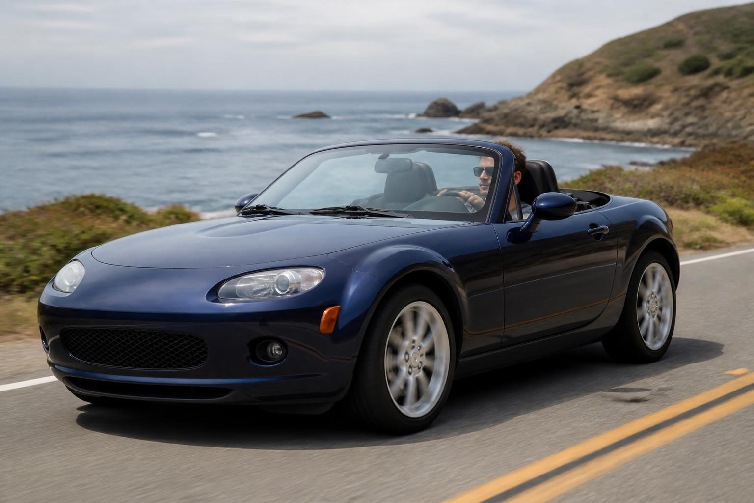 Blue two-seat convertible with roof down driving on a coastal road beside the ocean