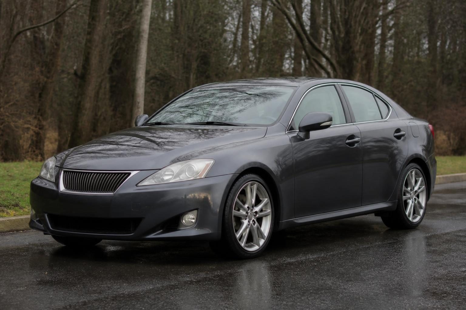 Gray Lexus IS four-door sedan parked on a wet road beside leafless trees.