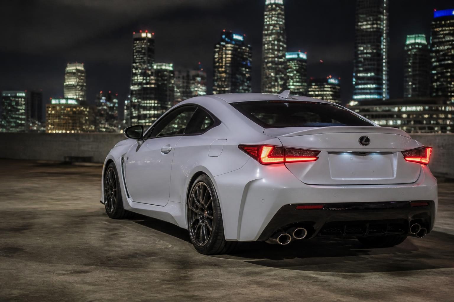 White Lexus RC F parked on a rooftop at night, viewed from the rear with city towers behind.