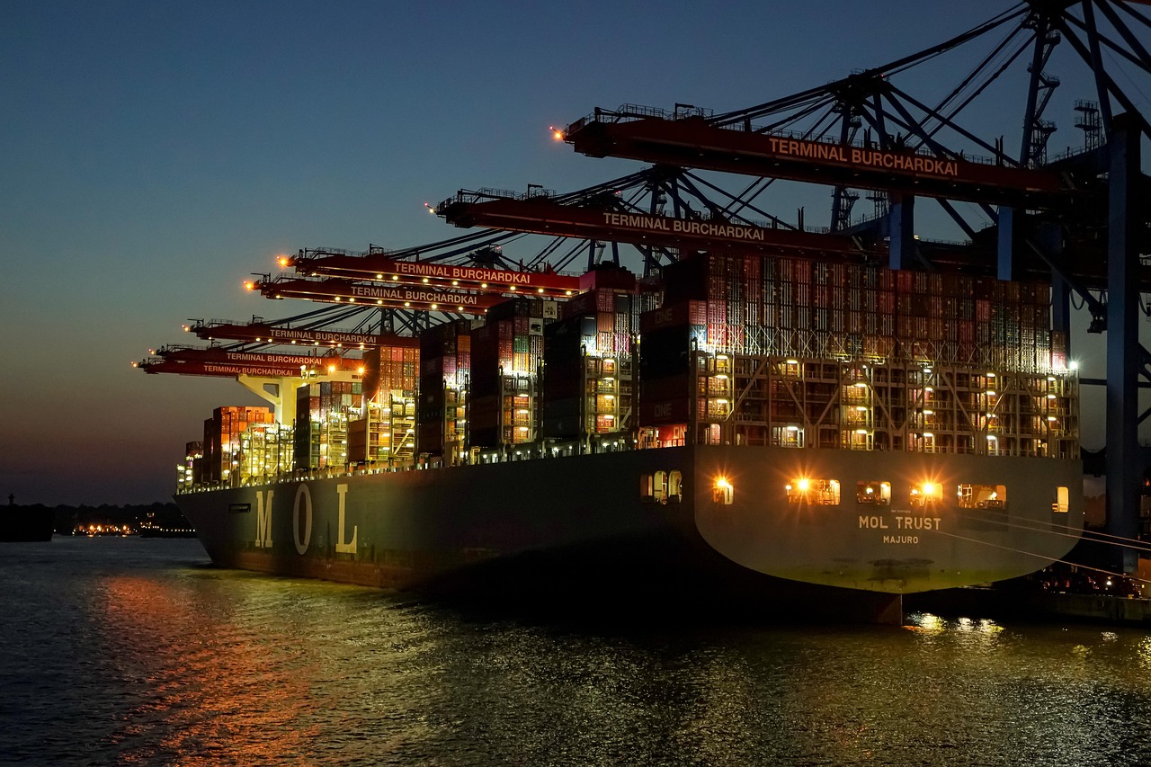 Container ship at a port at night under lit gantry cranes, with stacked containers and reflections on water