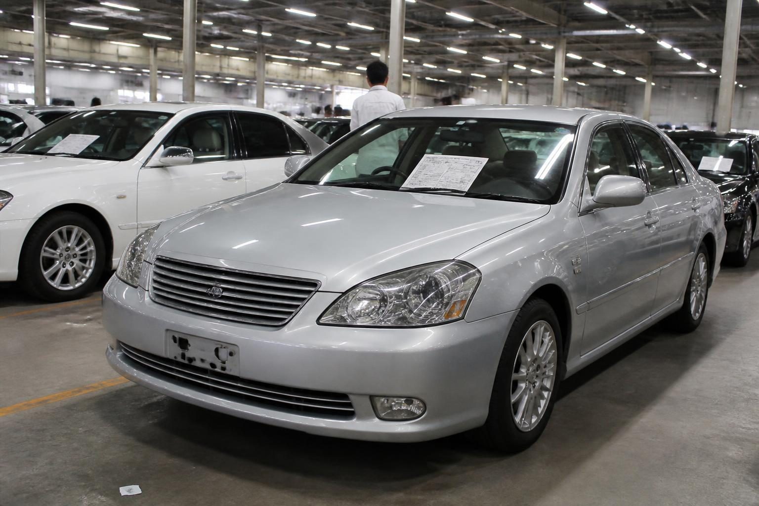 Silver sedan with a paper sheet on its windscreen parked among cars in a brightly lit indoor hall.