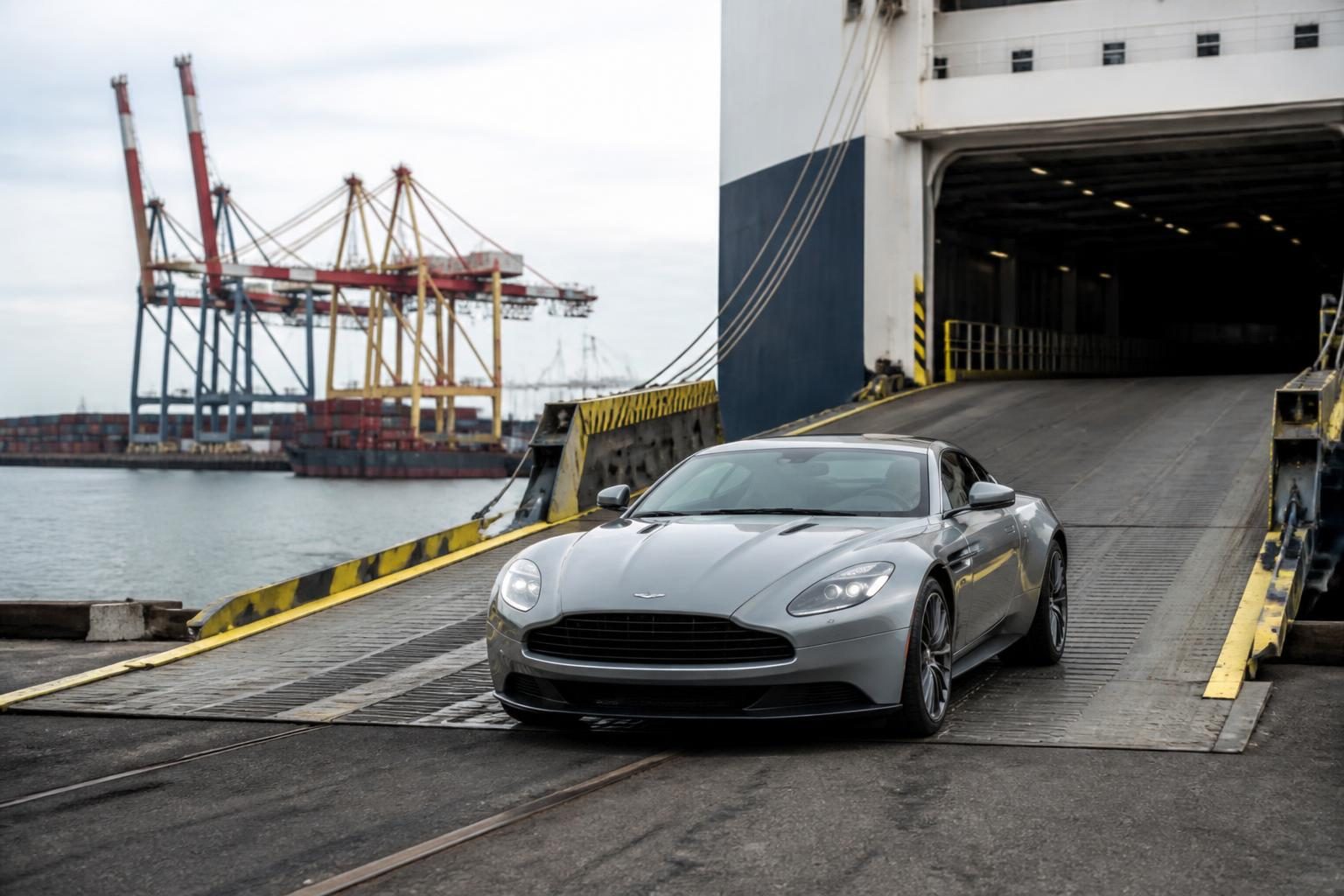 Silver sports car descending a cargo ship ramp beside cranes and containers at a port.