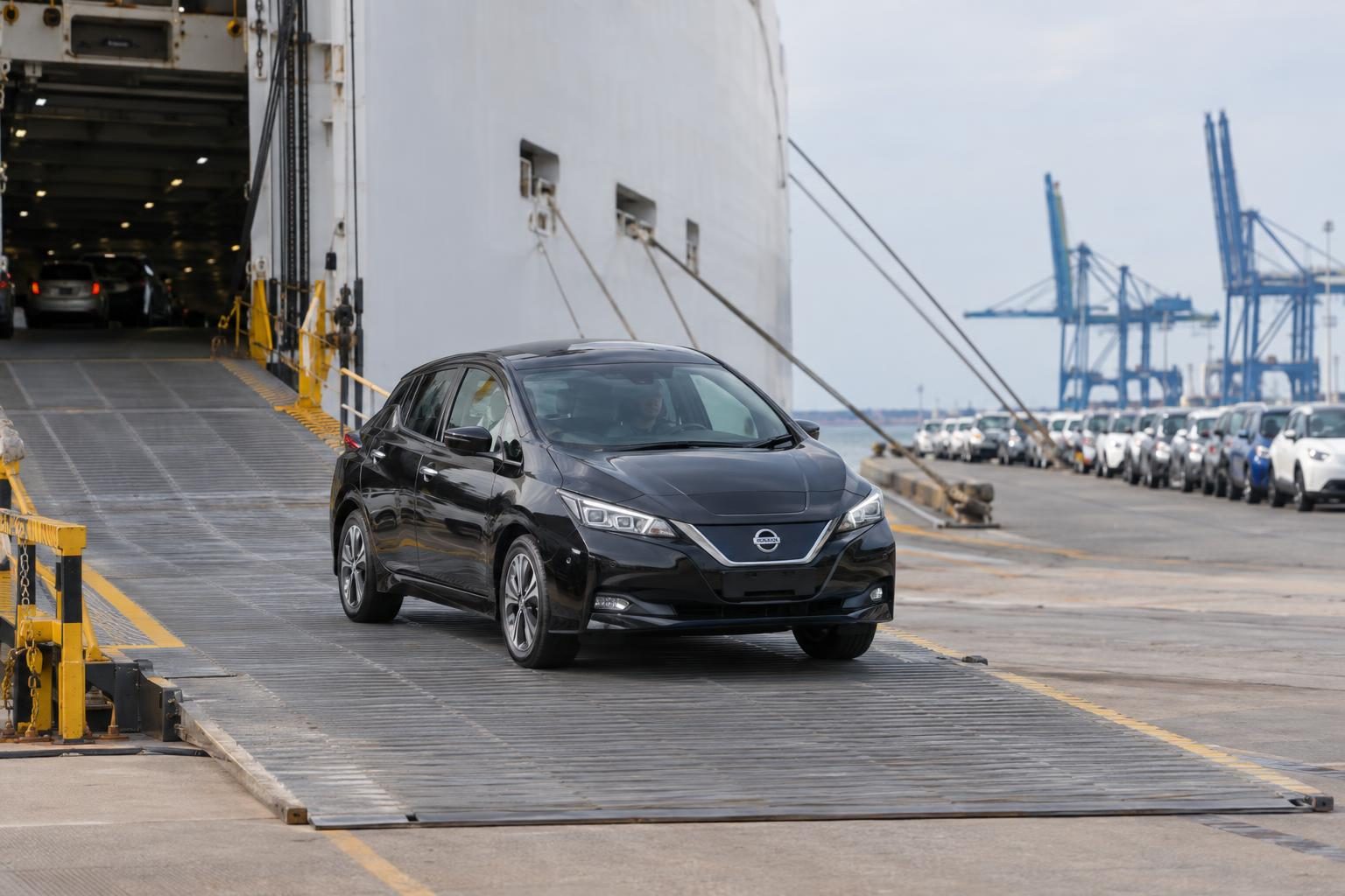 Black Nissan Leaf ZE1 on a RoRo vessel ramp at a port, with parked vehicles and cranes behind.