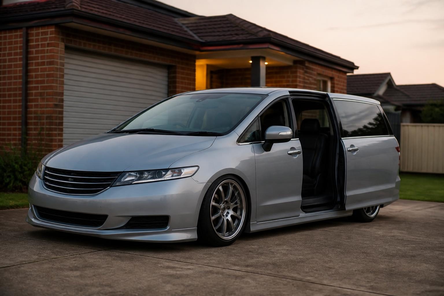 Silver minivan with open sliding door parked in front of a brick suburban house at dusk.