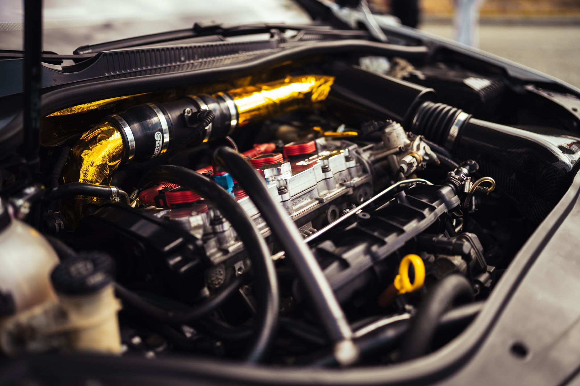 Close-up of a modified car engine bay with metal piping, red coil packs, and gold heat-wrapped tubing