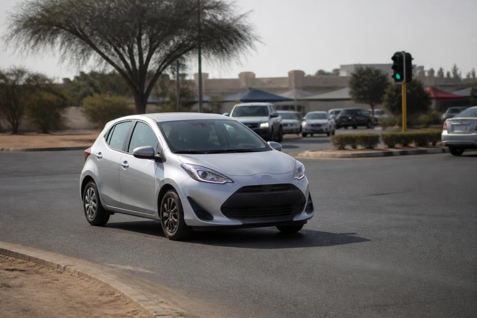 Silver compact hatchback driving through a suburban intersection with light traffic and dry trees
