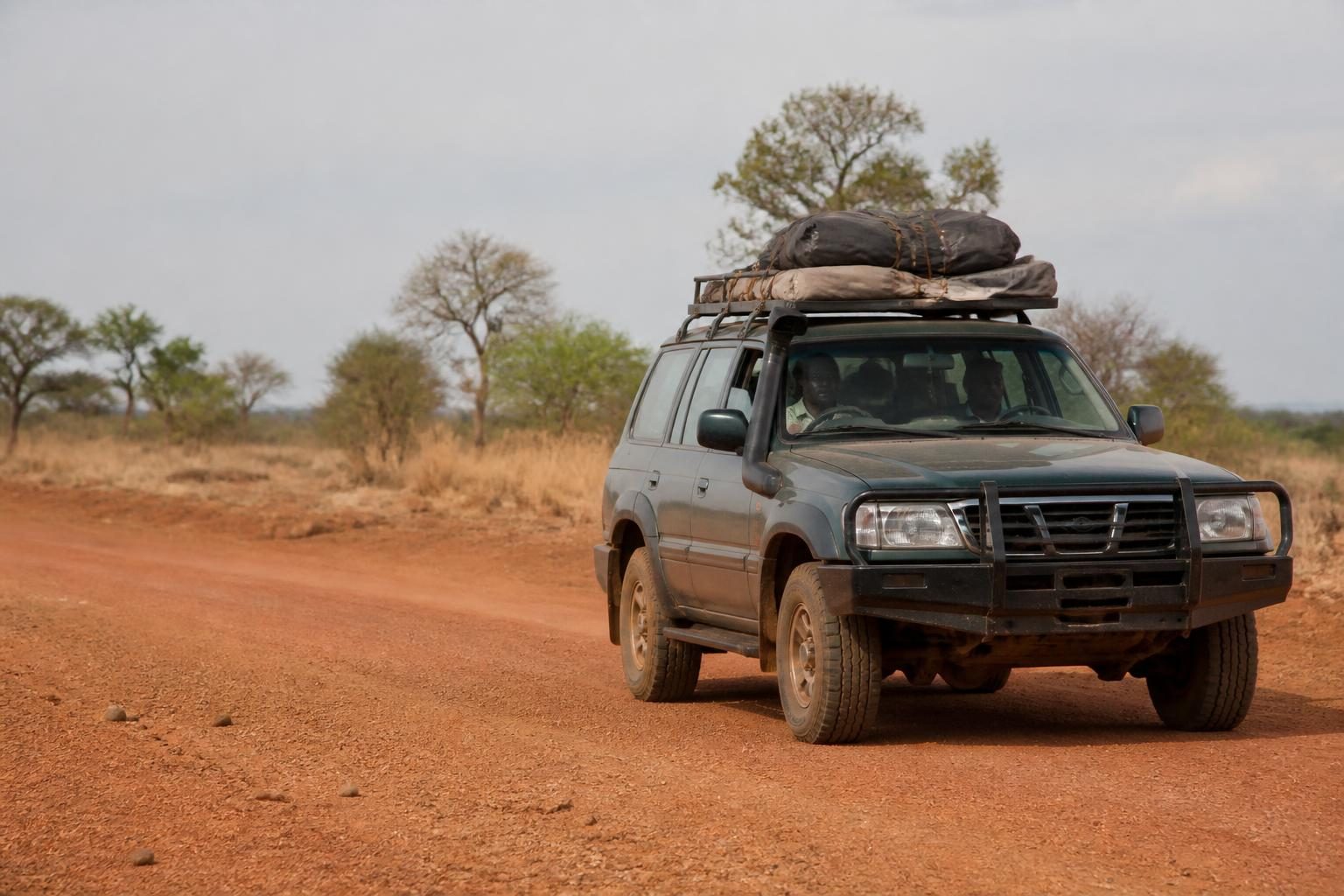 Rugged SUV with roof cargo driving on a red dirt road through dry savannah with scattered trees