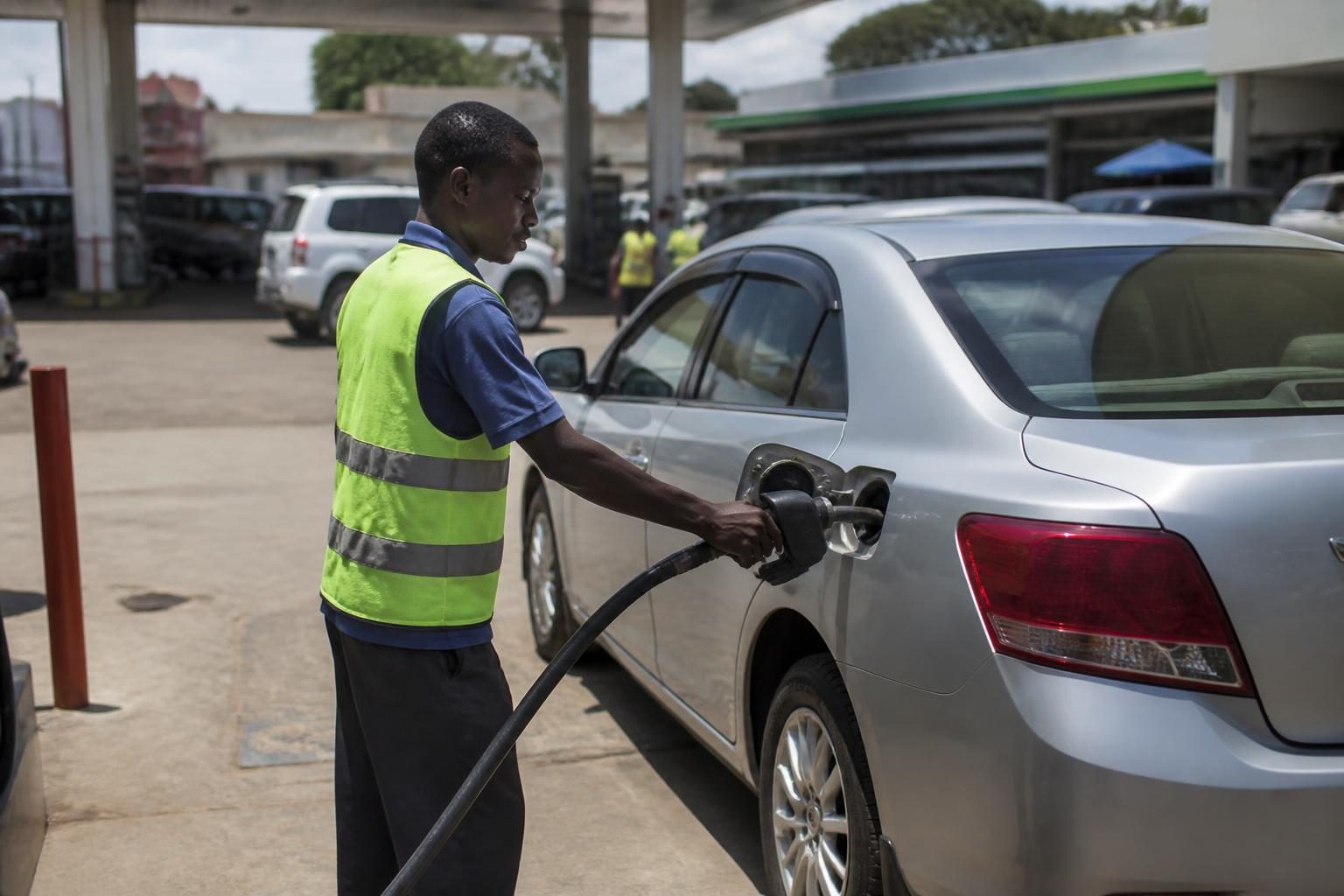 Fuel attendant in a yellow safety vest pumps fuel into a silver sedan at a busy station.