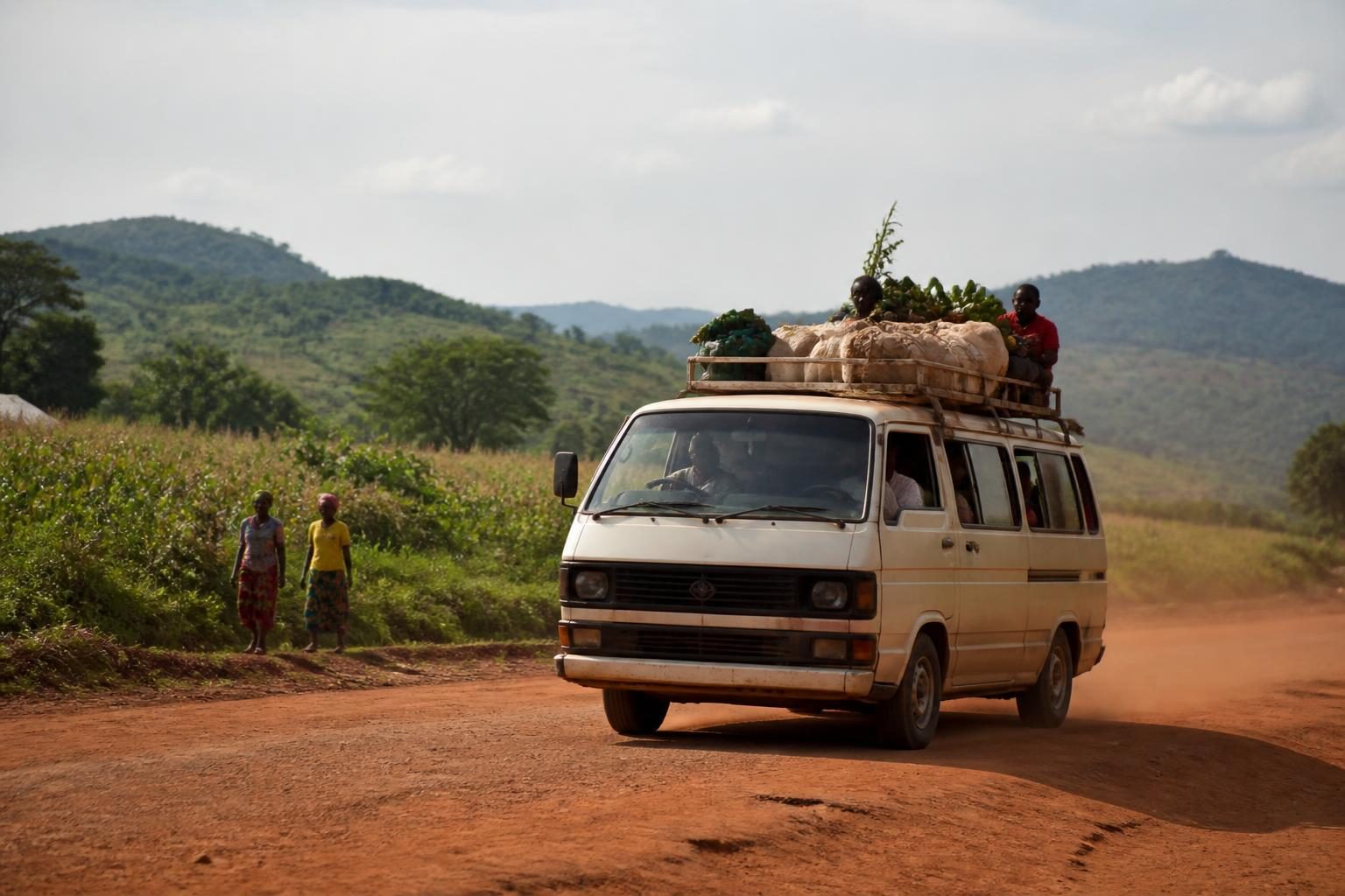 White minibus carrying sacks and produce on a red dirt road with hills and fields behind