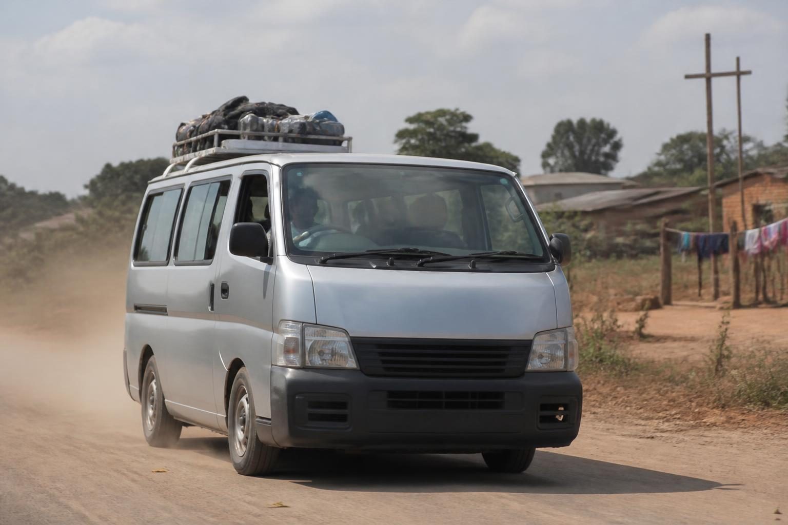 Silver passenger van with roof luggage driving on a dusty rural road beside small homes