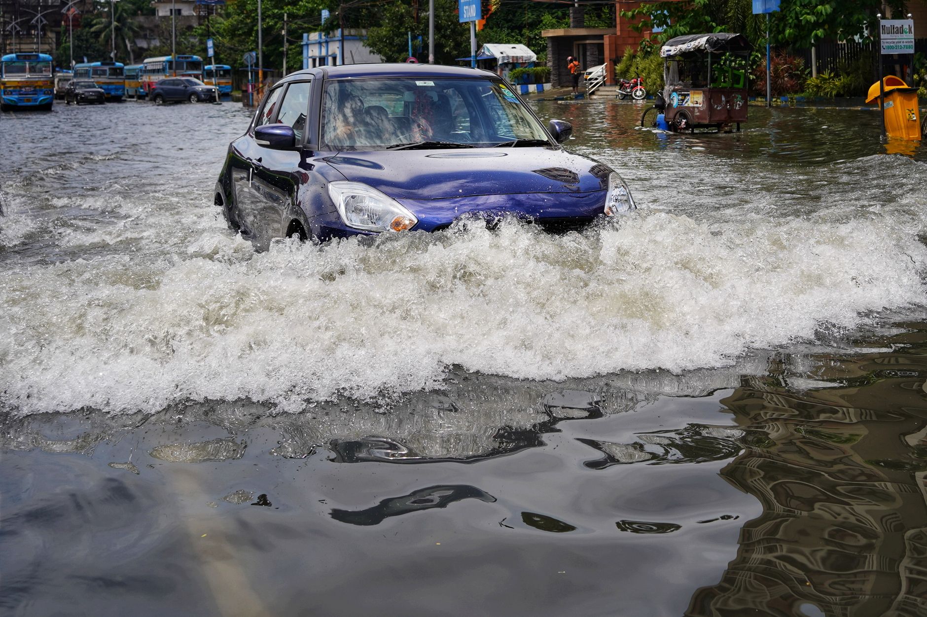 Blue car drives through floodwater on a city street, sending waves across the road.
