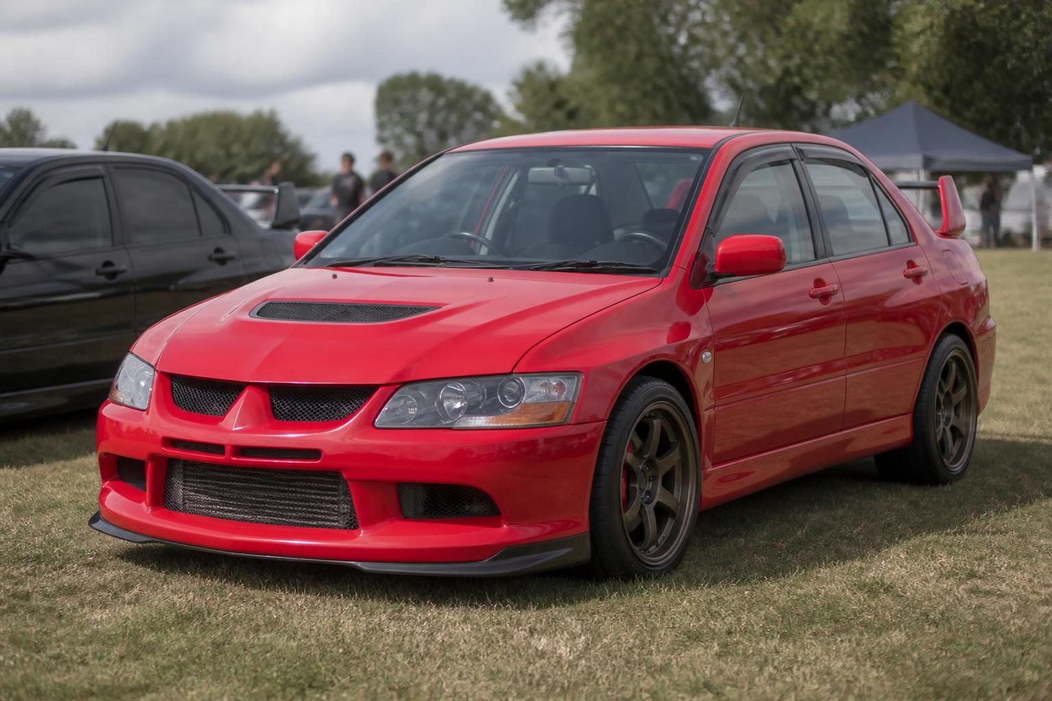 Red Mitsubishi Lancer Evolution with black wheels displayed on grass at an outdoor car show.
