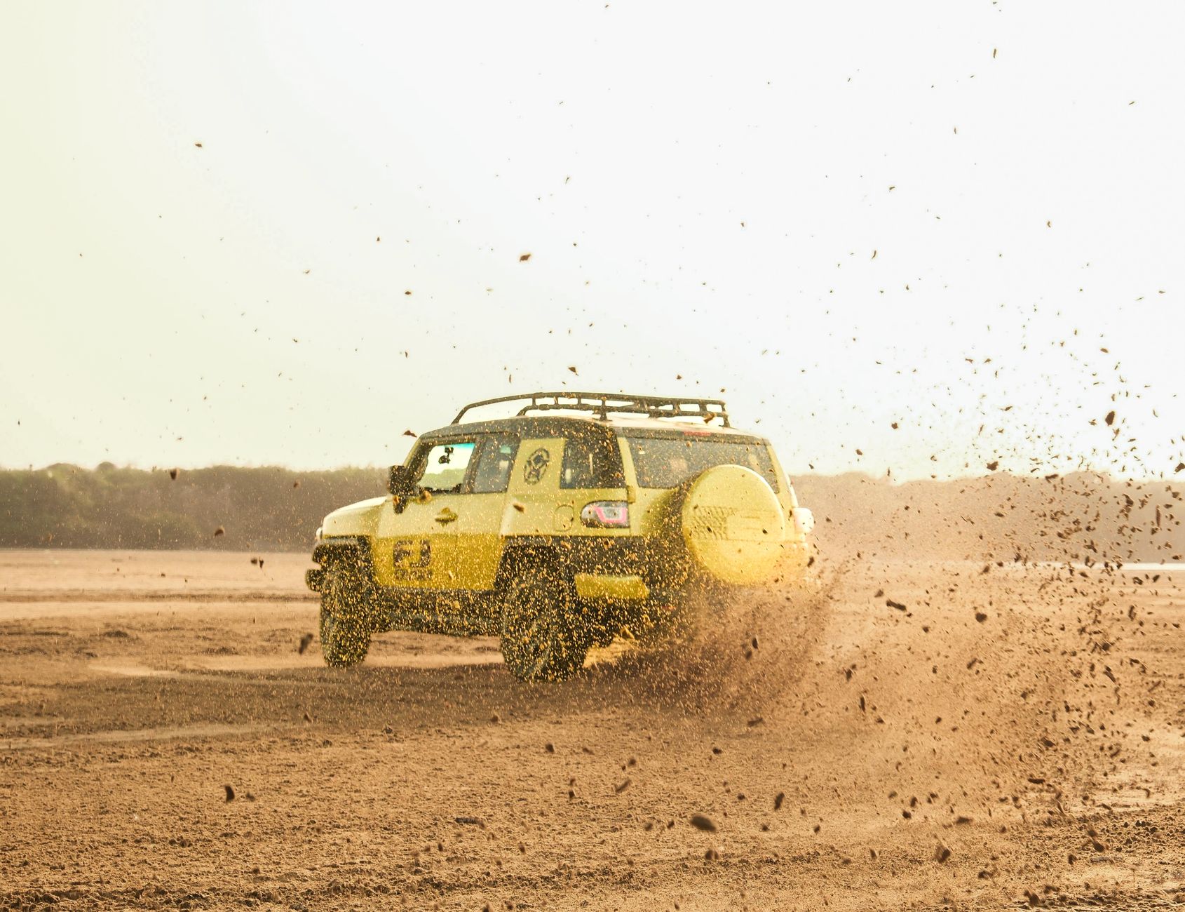 Yellow SUV driving across a dusty dirt track, spraying mud and gravel behind it.