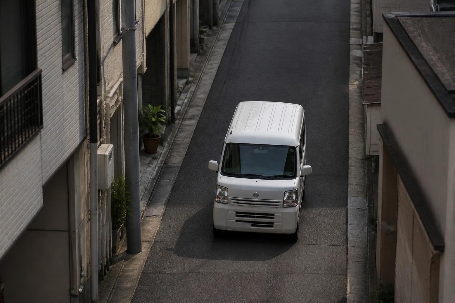 White compact kei van driving along a narrow street between closely packed buildings.