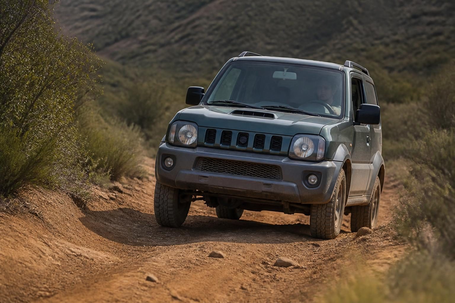 Silver Suzuki Jimny four-wheel drive climbing a rutted dirt track through scrubland.