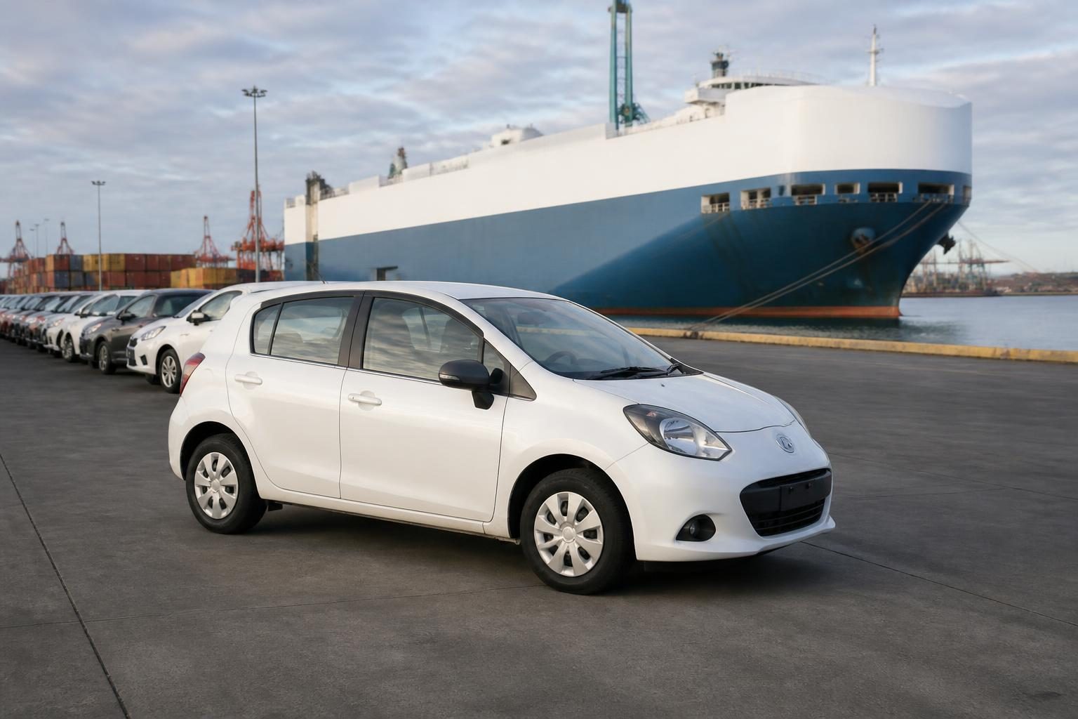 White compact cars parked on a wharf beside a blue-and-white car carrier ship.