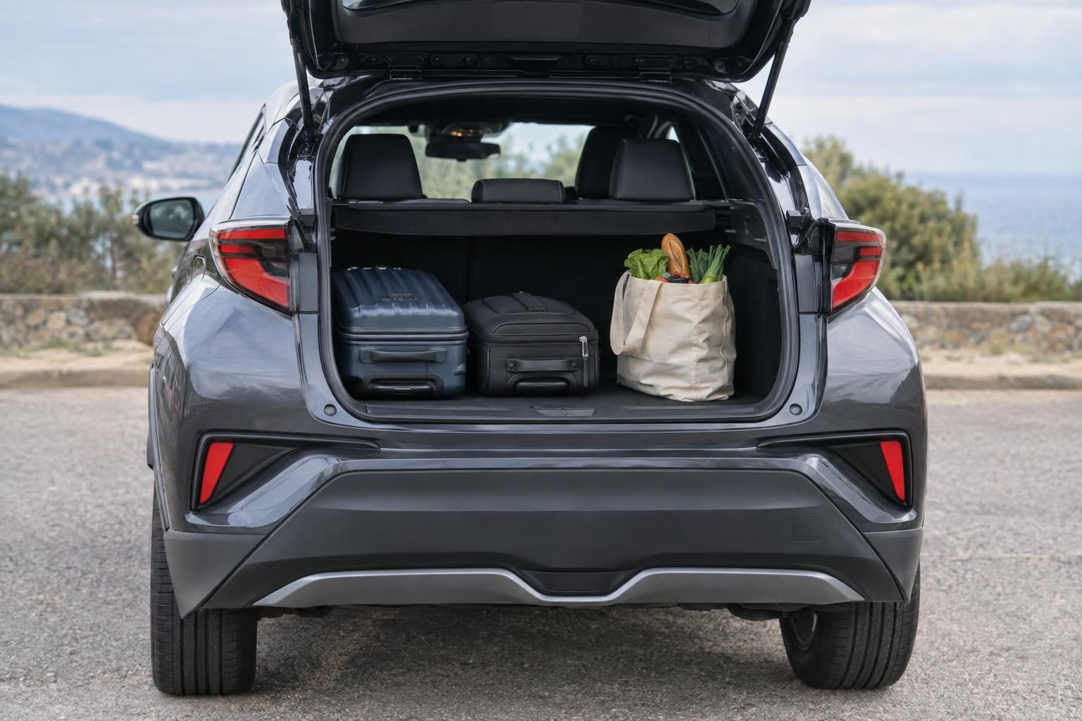 Open boot of a first-generation facelift Toyota C-HR with two suitcases and a grocery bag.