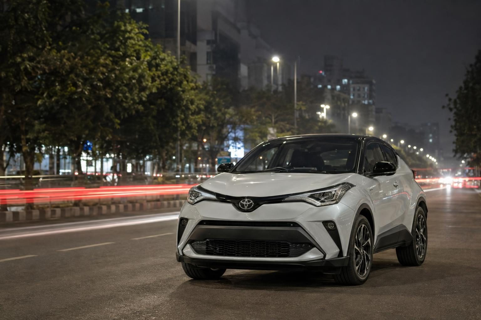 A white first-generation Toyota C-HR parked on a city street at night with traffic light trails.