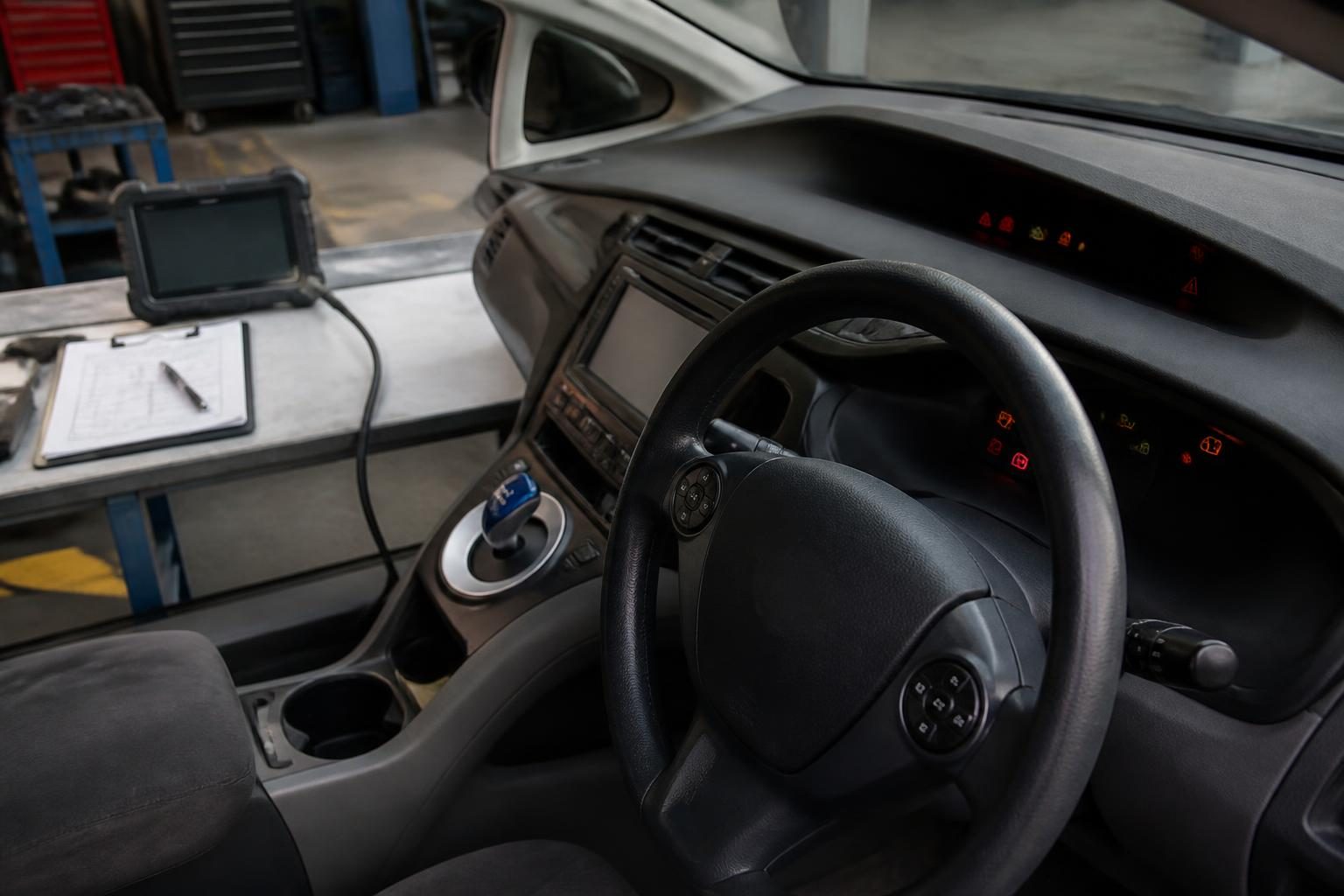 Right-hand-drive car dashboard with warning lights, diagnostic tablet, and inspection notes in a workshop.