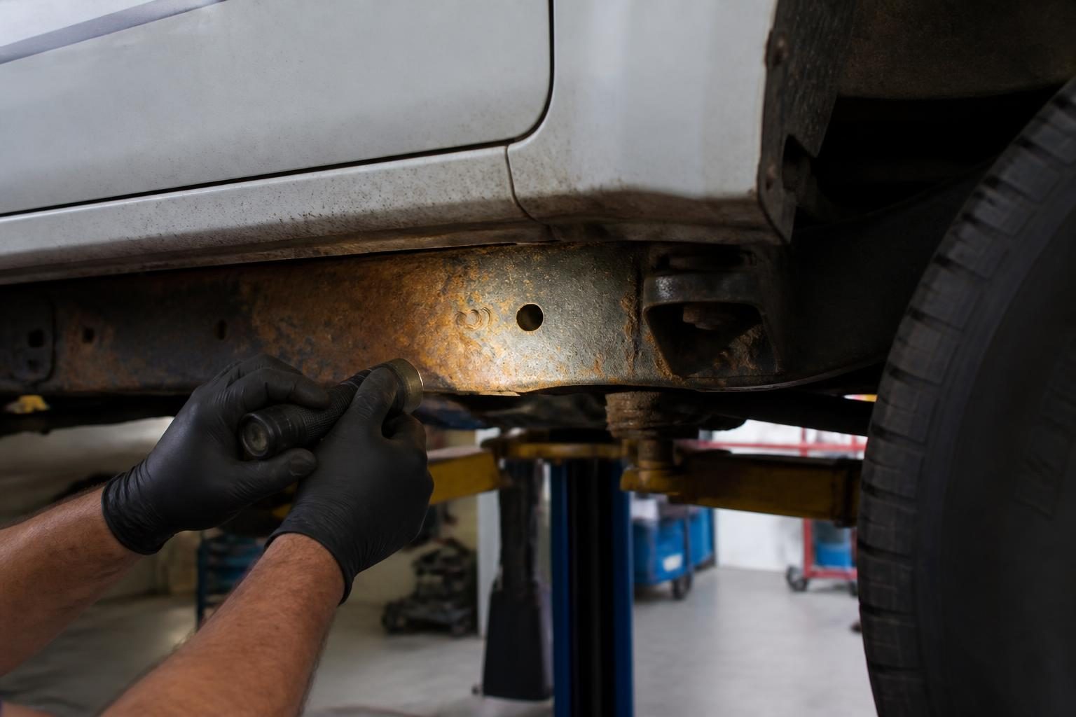 Gloved mechanic inspects a rusted chassis rail beneath a white vehicle on a lift.