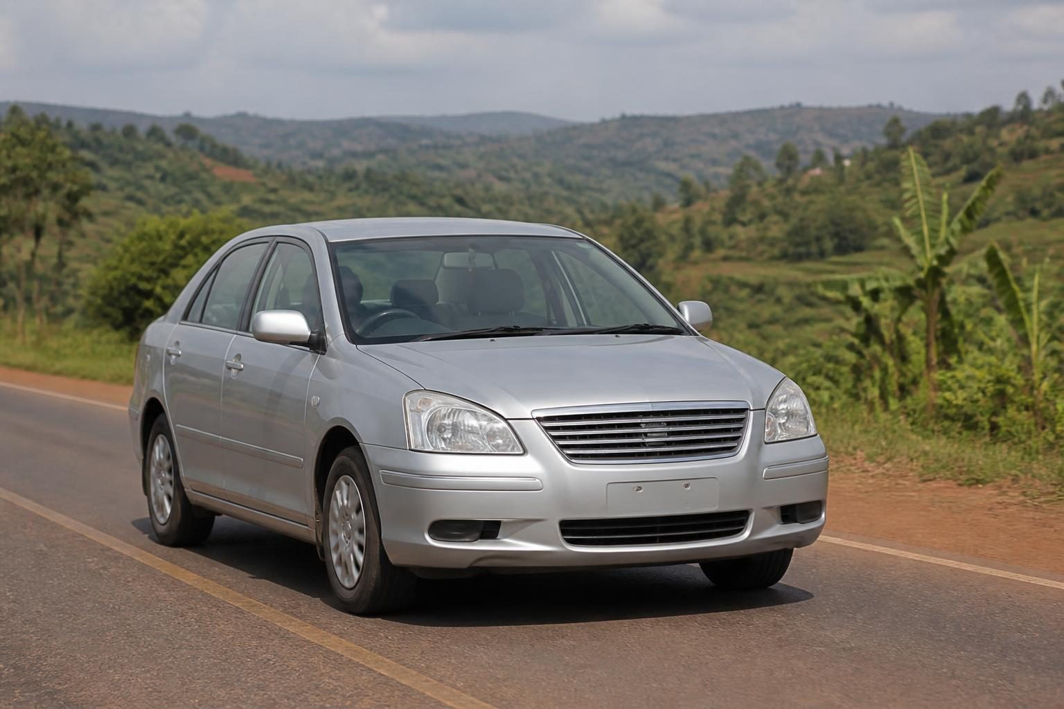 Silver four-door sedan driving on a paved rural road amid green hills and banana plants.