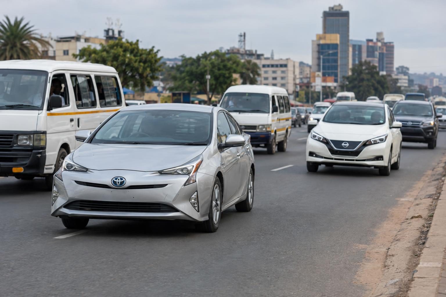 A Toyota Prius leads traffic beside minibuses and a Nissan Leaf on a busy city road.