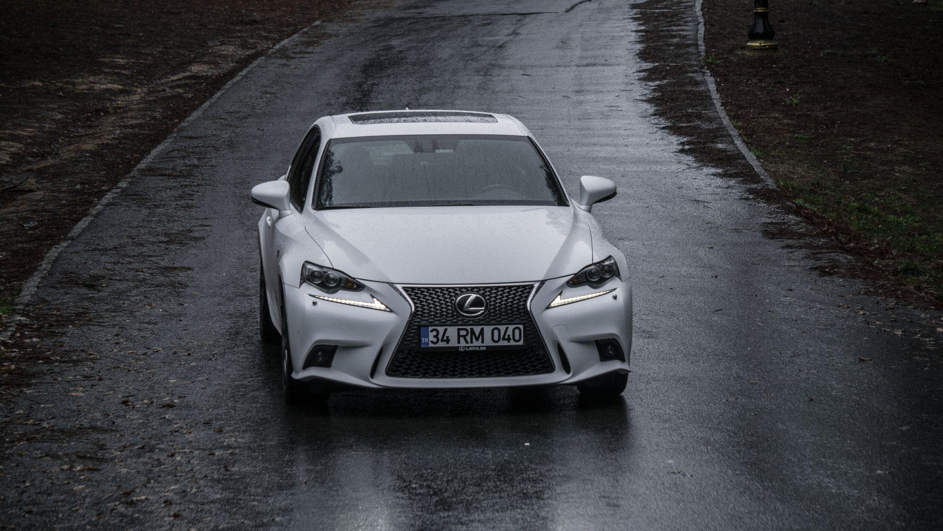 White Lexus IS sedan viewed from the front on a wet road with headlights on