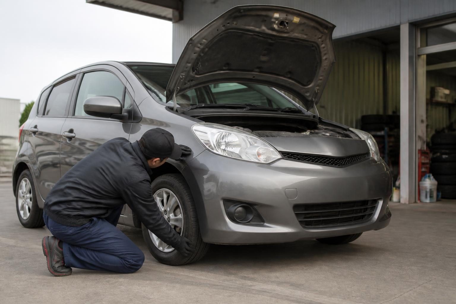 Mechanic kneels beside a grey hatchback, inspecting its front wheel while the bonnet is open.