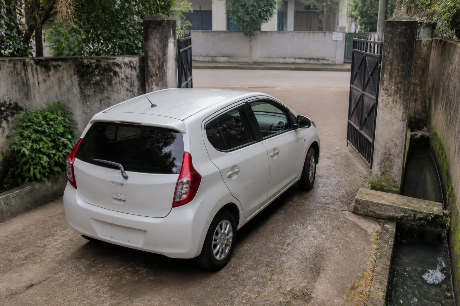 White compact hatchback at a narrow gated driveway beside an open concrete drain.