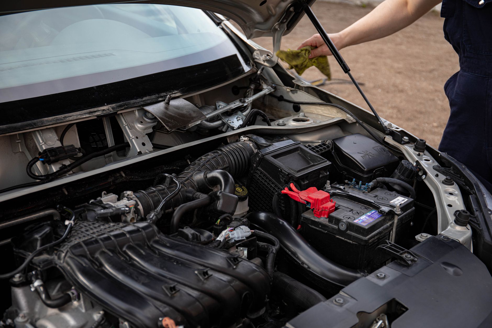 Open petrol-car engine bay with battery, air intake, fluid reservoir and a person inspecting it.